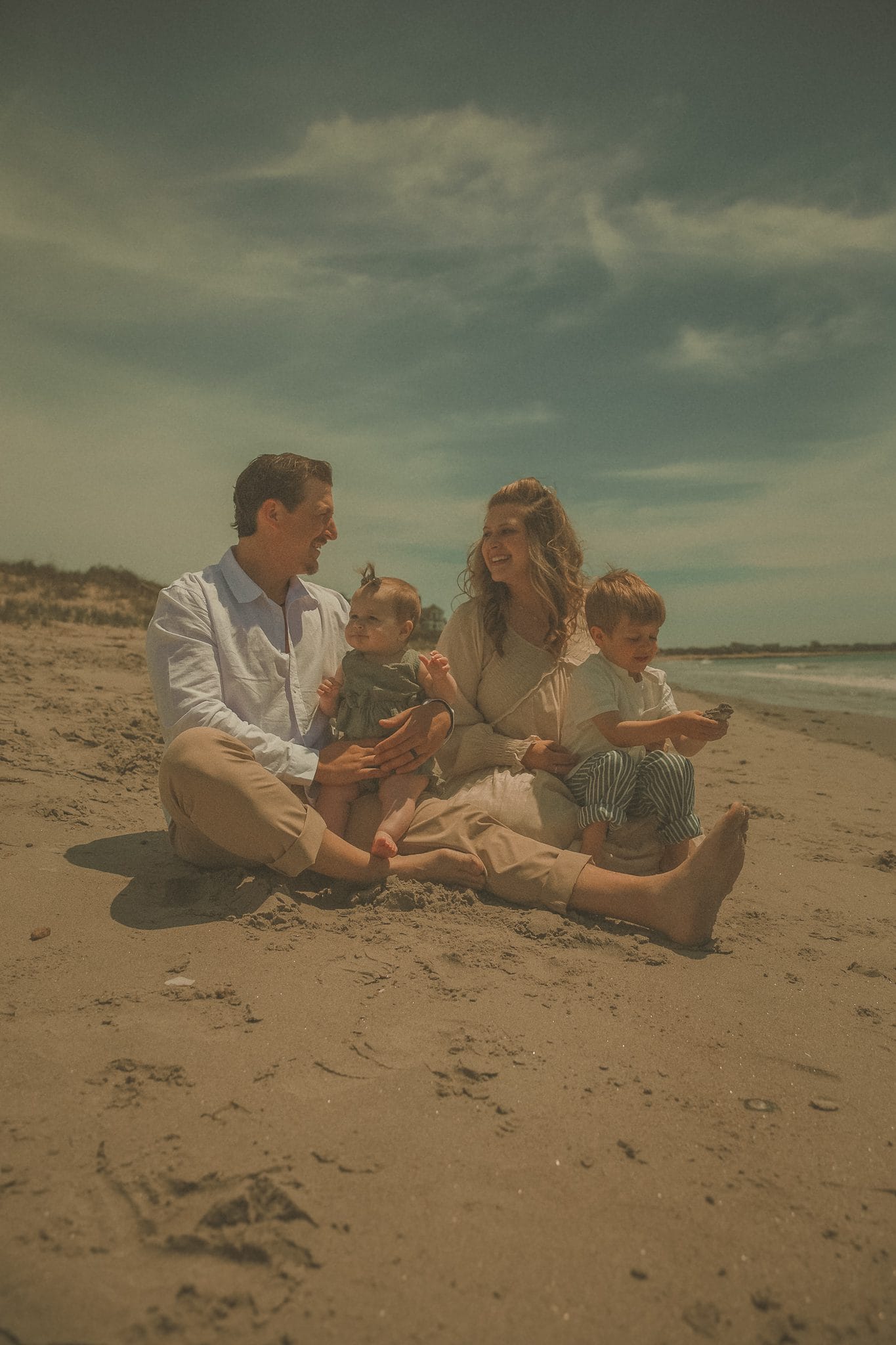 Family of four sitting together on a sandy beach, parents smiling at each other while holding a baby and toddler on a sunny day