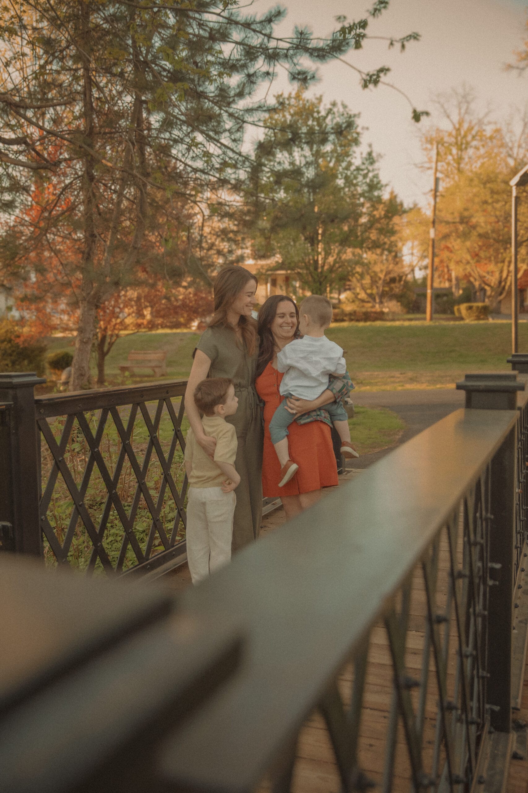 Two mothers smiling together on a wooden bridge with their young children during an autumn family portrait session in Connecticut at golden hour