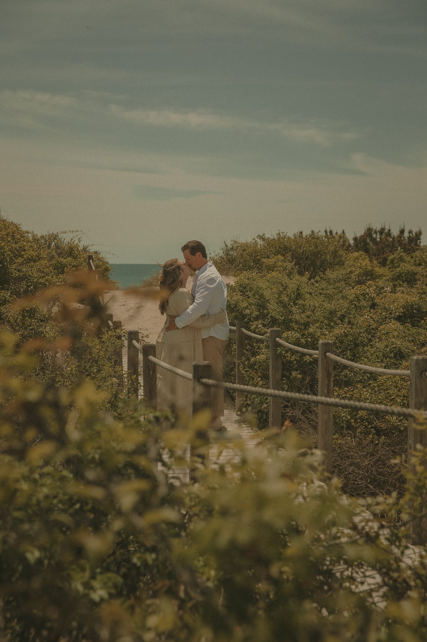 Connecticut couples photographer captures intimate kiss on a weathered boardwalk surrounded by lush coastal greenery and ocean beyond
