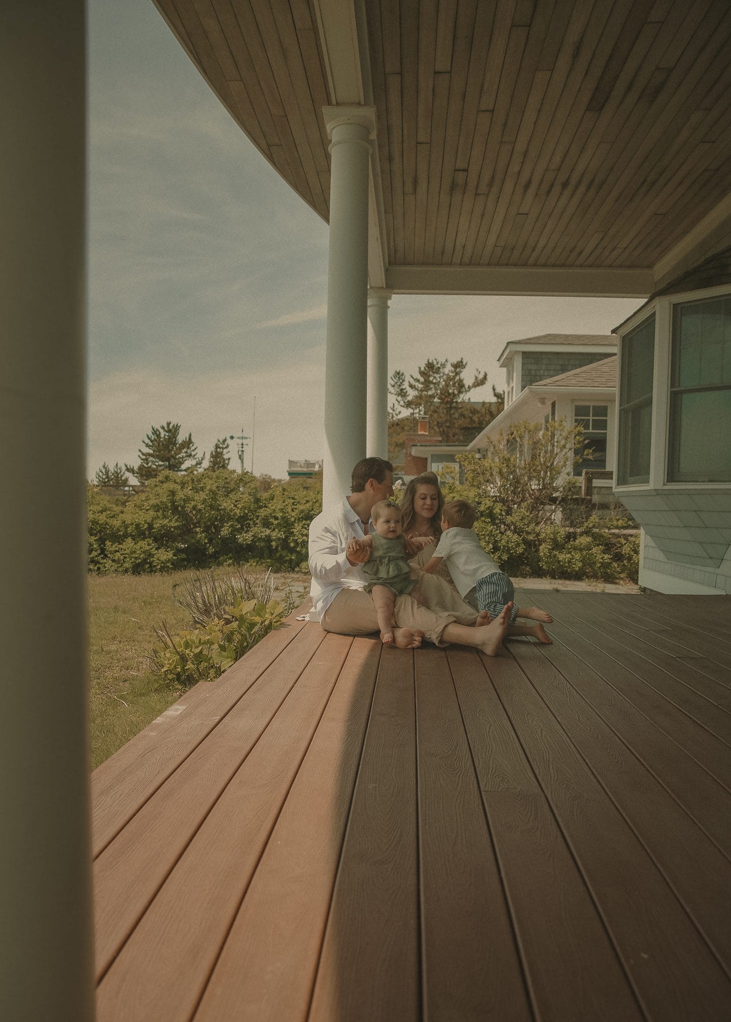 Connecticut family photographer captures young family of four sitting together on a sun-warmed coastal porch with ocean greenery behind them