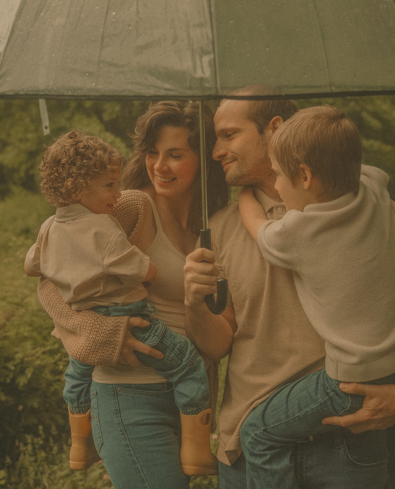 Connecticut family photographer captures a family of four laughing together under a clear umbrella during an outdoor rainy day session in lush green surroundings