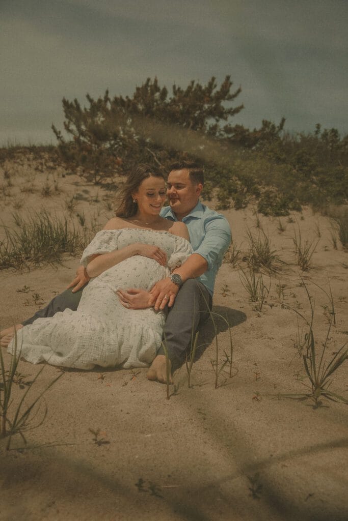 Connecticut maternity photographer captures expecting couple seated together in coastal sand dunes, hands resting on baby bump, surrounded by beach grass