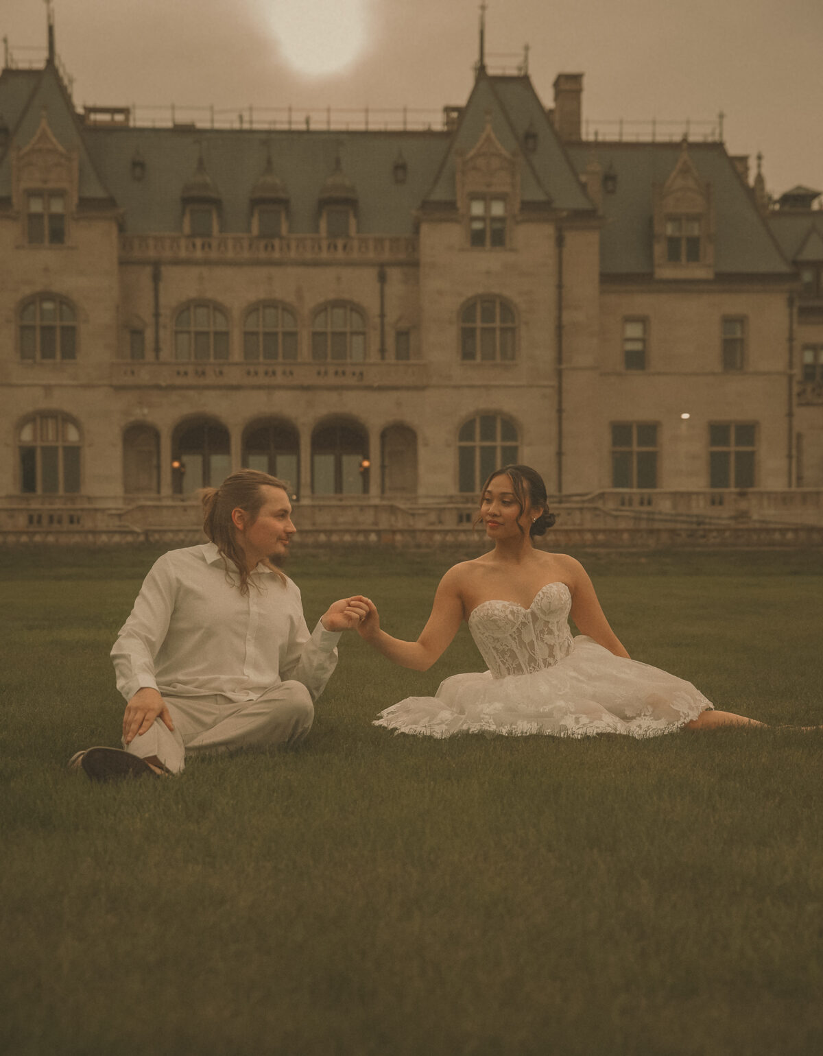 Connecticut wedding photographer captures bride and groom sitting together on the lawn holding hands in front of a grand stone estate under a dramatic evening sky