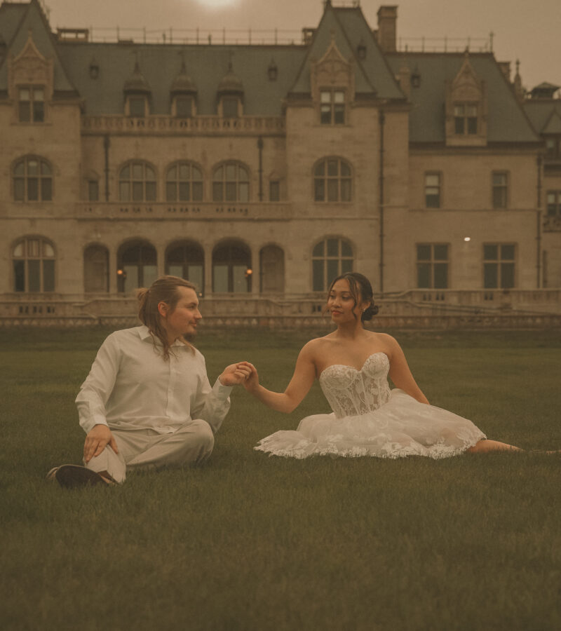 Connecticut wedding photographer captures bride and groom sitting together on the lawn holding hands in front of a grand stone estate under a dramatic evening sky