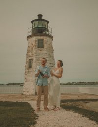 Couple laughing and popping champagne in front of a historic stone lighthouse with waterfront views in the background
