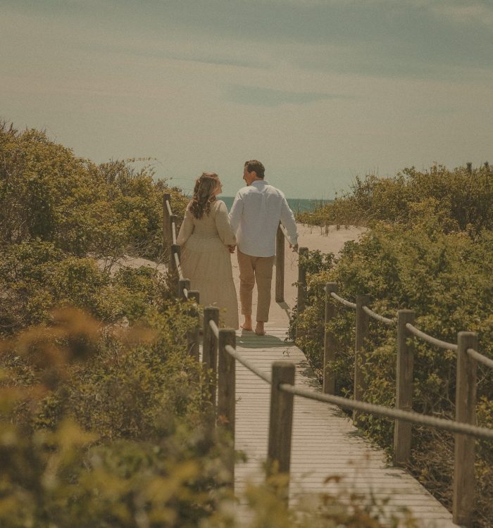 Connecticut couples photographer captures pair walking hand in hand down a wooden boardwalk path through coastal dunes toward the ocean