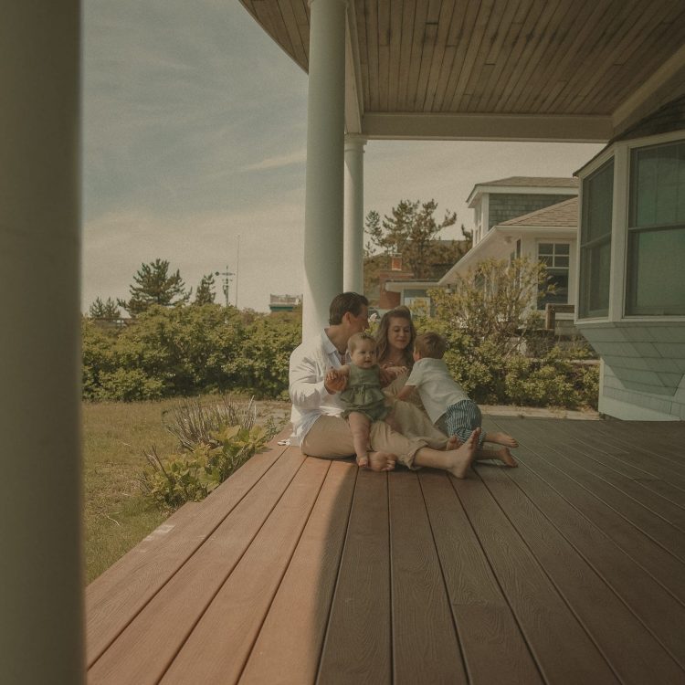 Connecticut family photographer captures young family of four sitting together on a sun-warmed coastal porch with ocean greenery behind them