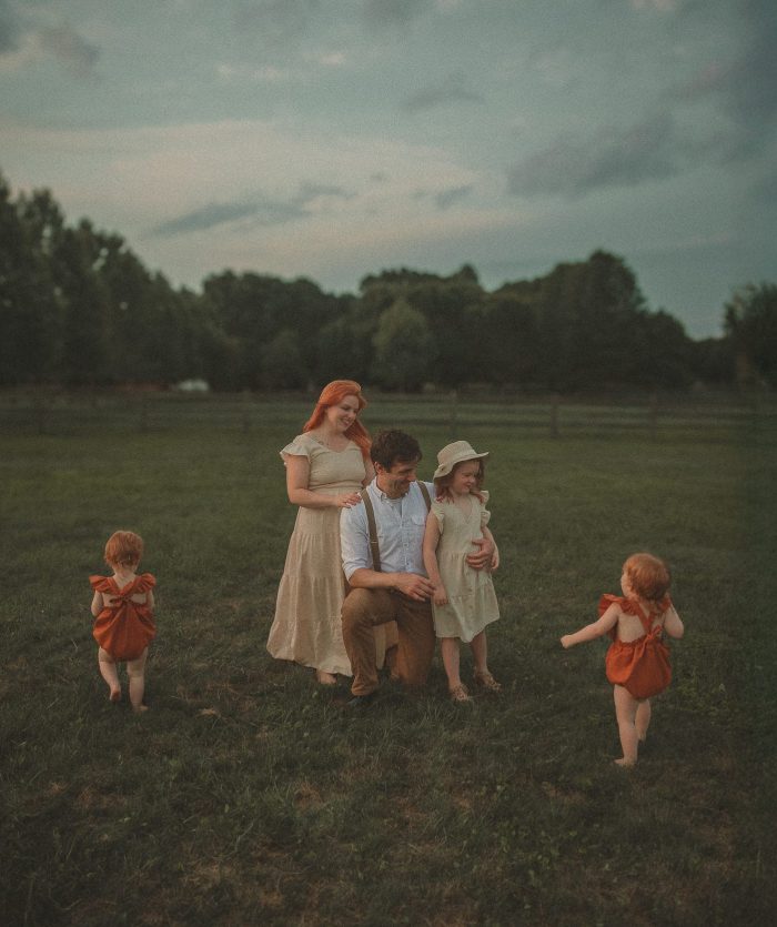 Connecticut family photographer captures a candid golden hour portrait of a family of five in an open field. A red-haired mother in a cream dress and father in suspenders kneel with their older daughter while twin red-haired toddlers in rust rompers play freely around them under a dramatic evening sky.
