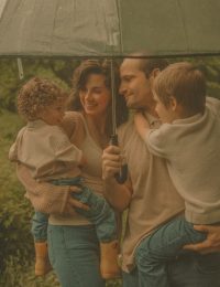 Connecticut family photographer captures a family of four laughing together under a clear umbrella during an outdoor rainy day session in lush green surroundings
