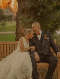 Connecticut wedding photographer captures bride and groom seated on a wooden bench beneath an autumn tree, sharing a quiet and tender moment