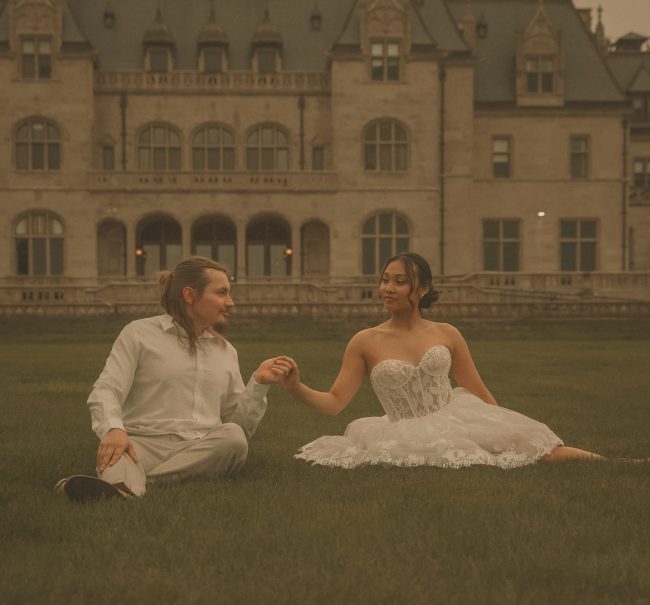 Connecticut wedding photographer captures bride and groom sitting together on the lawn holding hands in front of a grand stone estate under a dramatic evening sky