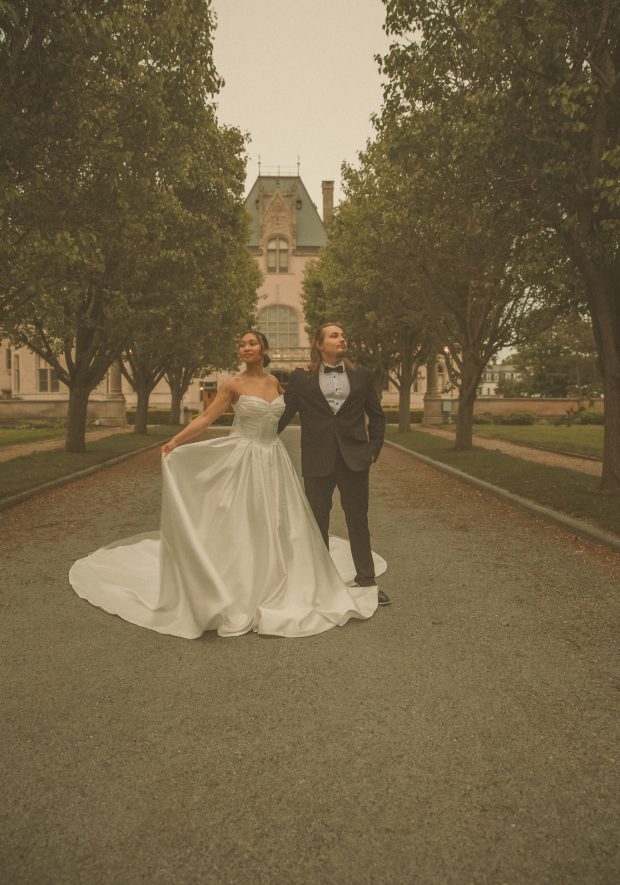 Connecticut wedding photographer captures bride in a sweeping ball gown and groom in a tuxedo standing back to back on a tree-lined path in front of a historic estate