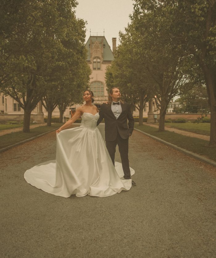 Connecticut wedding photographer captures bride in a sweeping ball gown and groom in a tuxedo standing back to back on a tree-lined path in front of a historic estate