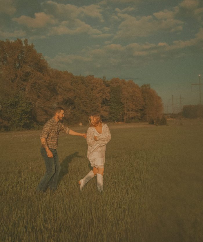 Couple dancing and laughing together in an open green field at golden hour, with trees and a cloudy sky in the background
