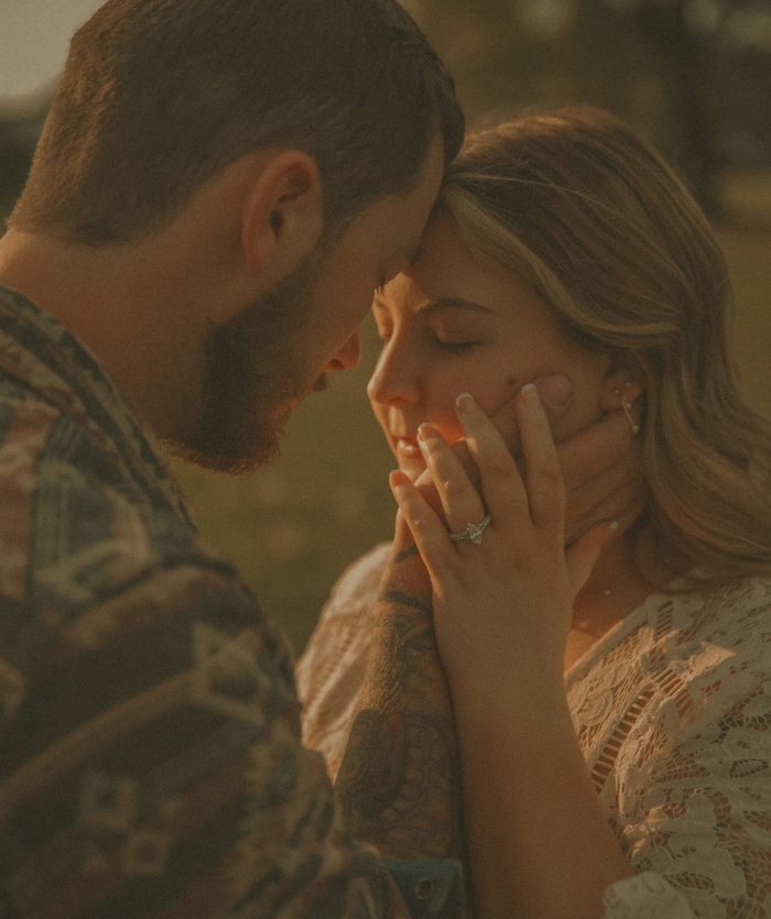 Intimate close-up of an engaged couple with foreheads touching, the woman's hand showing an engagement ring, bathed in warm sunset light